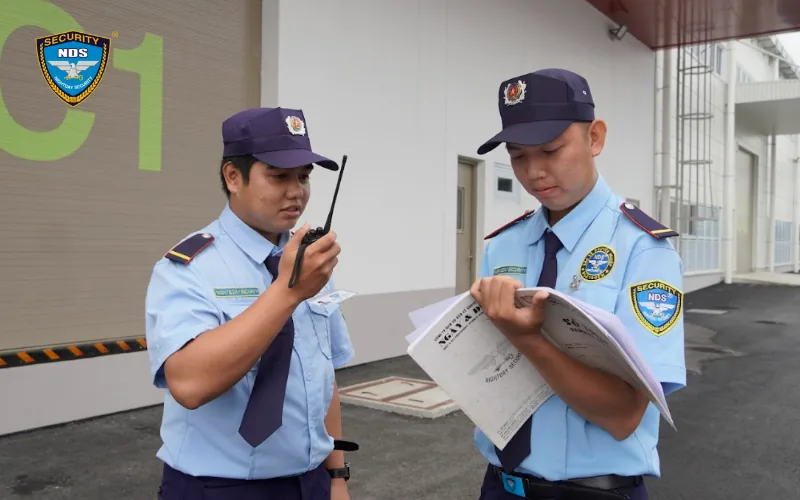 Hai nhân viên bảo vệ Ngày & Đêm dùng bộ đàm và ghi sổ trực tại cửa nhà kho