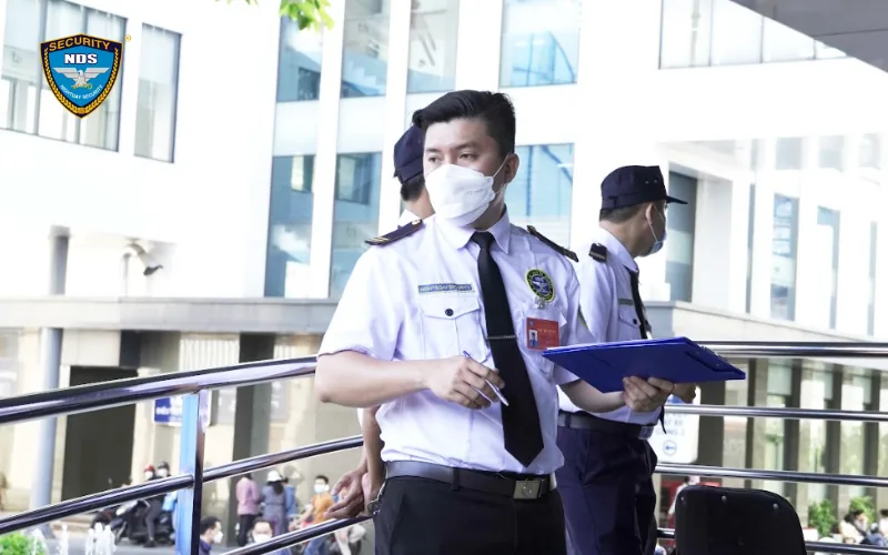 Nhân viên bảo vệ Ngày & Đêm đeo khẩu trang, cầm bìa hồ sơ tại lối ra vào